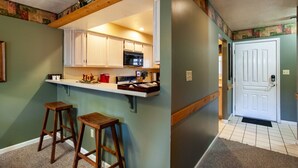 Cocina con armarios blancos, microondas y encimera con varios objetos. Hay dos taburetes de madera y una puerta que da a otra habitación.