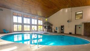Una piscina interior con paredes de vidrio transparente, techo de madera y algunas sillas.