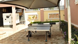 Ein Tischtennis-Tisch im Freien unter einem großen Sonnenschirm, umgeben von einer gepflasterten Terrasse, mit einem Gebäude im Hintergrund.
