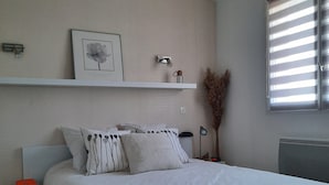 Un dormitorio con una cama grande, una mesita de noche, un estante en la pared con una imagen y un objeto pequeño, un radiador y una ventana con persianas.