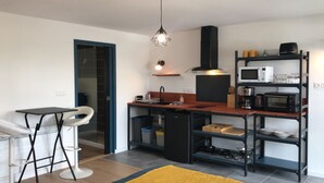 A compact kitchen with a dining area, a black refrigerator, a microwave, a coffee maker, and a shelf with kitchenware. - Studio 35 m2 Refait à Neuf (Fougères)