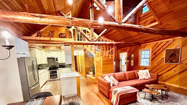 A rustic cabin interior with a wooden dining table, a sofa, and a kitchen area with a refrigerator and stove.