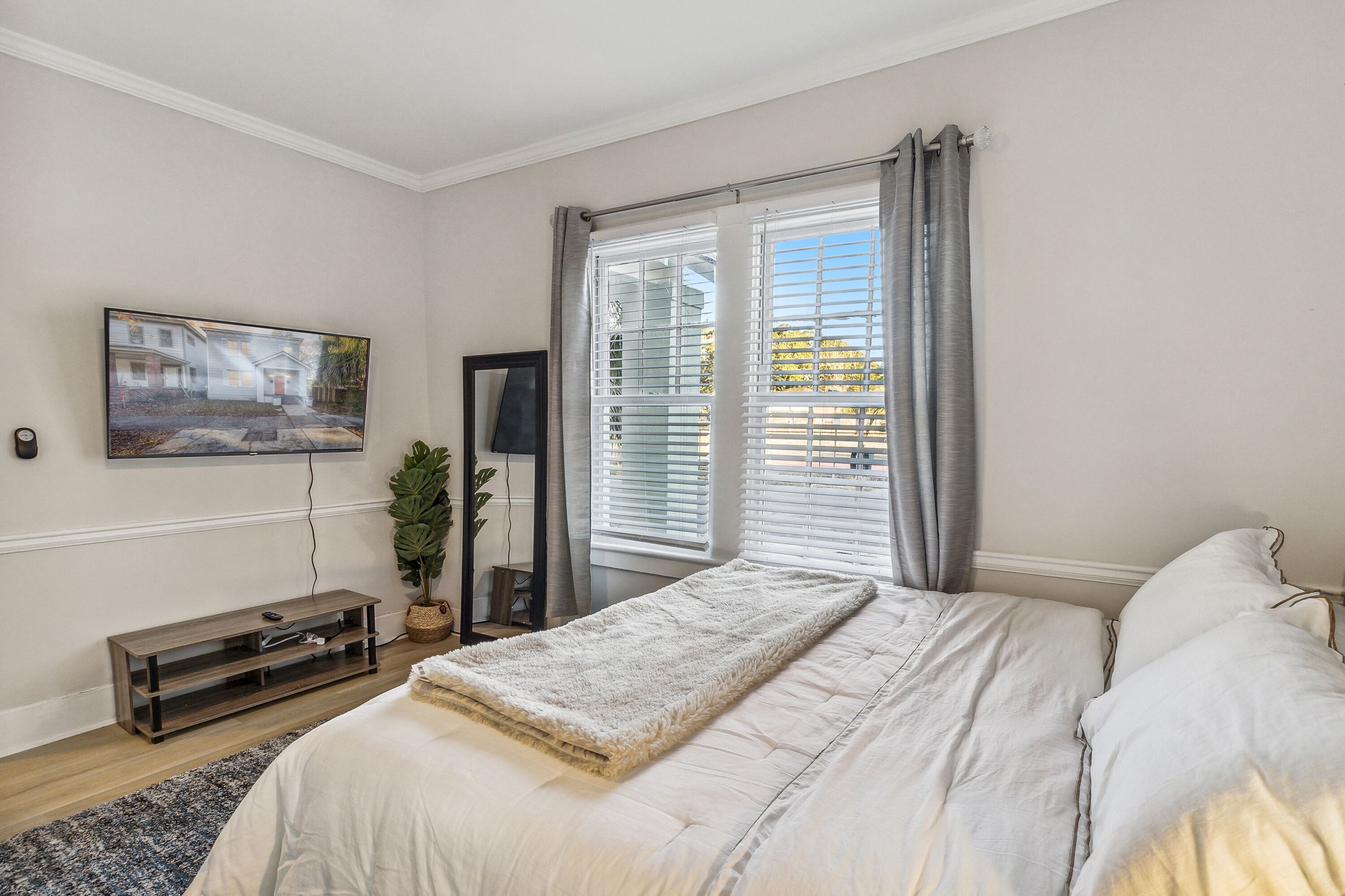 Un dormitorio con cama, televisor en la pared, ventana con cortinas y una planta.