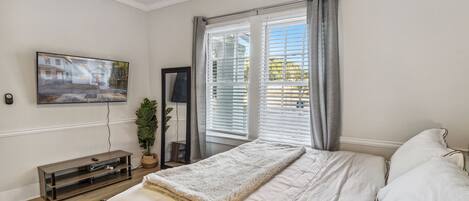 Un dormitorio con cama, televisor en la pared, ventana con cortinas y una planta.