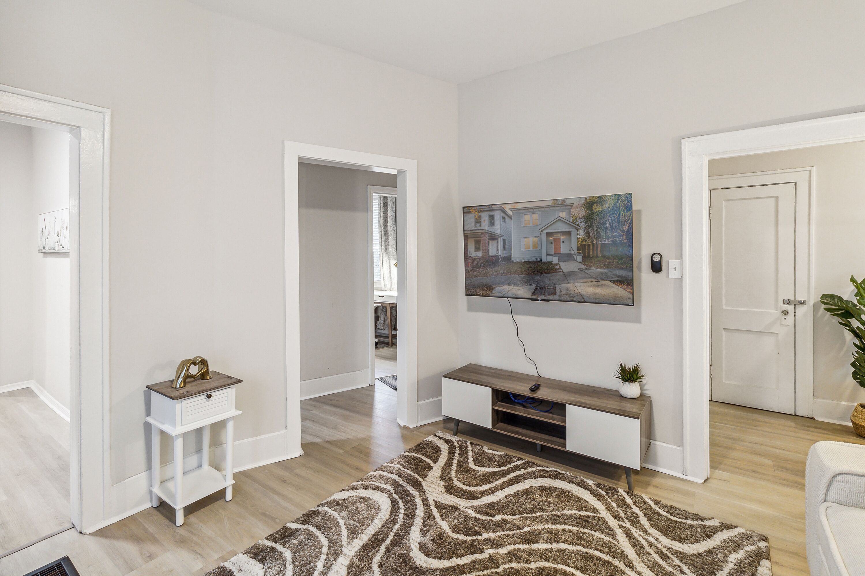 Una sala moderna con un televisor de pantalla plana montado en la pared, una mesa consola de madera, una alfombra estampada y una puerta que da a otra habitación.