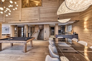 A spacious room with a pool table, a dining area with a long table and chairs, wooden walls, and a staircase leading to an upper level.