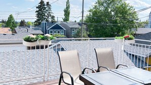 Terraza en la azotea con una mesa y sillas, rodeada por una cerca blanca, con vistas a una zona residencial con casas y árboles.