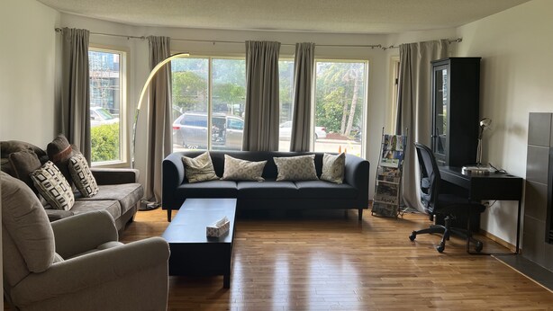 Sala de estar con sofá, sillón y mesa de centro. Se ve un mueble para televisión con televisor y teclado. Amplias ventanas con cortinas.