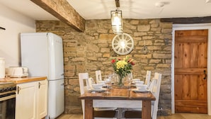 Un coin repas avec une table en bois dressée pour six personnes, entourée de chaises blanches, d’un mur en pierre et d’une porte en bois.