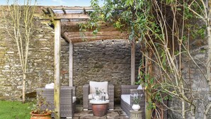 Un jardin clos par un mur de pierres, agrémenté de meubles en osier, de plantes en pot et d’une pergola rustique.