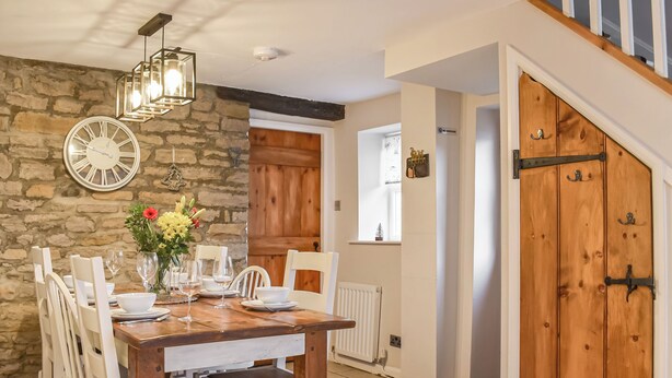 Un coin repas avec une table en bois dressée pour quatre personnes, des chaises blanches, un mur en pierre, une horloge et un luminaire suspendu.
