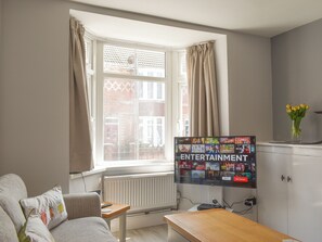 Uma sala de estar com sofá, mesa de centro, TV fixada na parede exibindo diversas opções de entretenimento, janela com cortinas e um vaso de flores em um aparador.