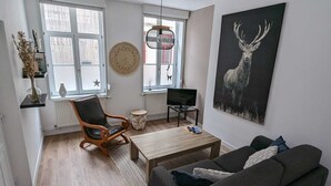 A modern living room with a wooden floor, a black leather chair, a grey sofa, a wooden coffee table, a TV on a stand, and a large deer painting on the wall. - En Hypercentre, au Calme, Avec Cour Privative Aménagée, Appartement Rénové (Arras)