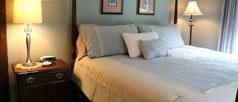 Een net opgemaakt bed met een witte deken en meerdere kussens, een nachtkastje met een lamp, en twee ingelijste foto's aan de muur.