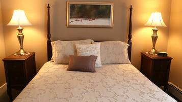 A neatly made bed with a patterned bedspread, flanked by two bedside tables with lamps, a framed picture above the bed, and a nightstand with a clock.