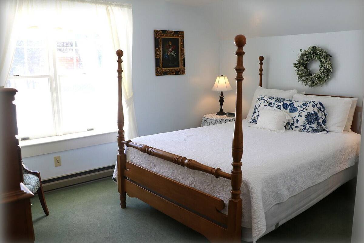 Ein Bett mit Baldachin, mit weißer Bettdecke, ein Nachttisch mit Lampe, ein gerahmtes Bild an der Wand und ein Kranz über dem Bett.