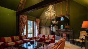 A spacious living room with a high ceiling, exposed wooden beams, a large chandelier, and a cozy seating area with patterned upholstery.