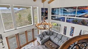 A modern living room with a gray sectional sofa, a wooden coffee table, and a chandelier. Large windows offer a view of a mountainous landscape. - Mountainside Chateau spectacular views and sunsets. 15 min to downtown Ashland! (Ashland)