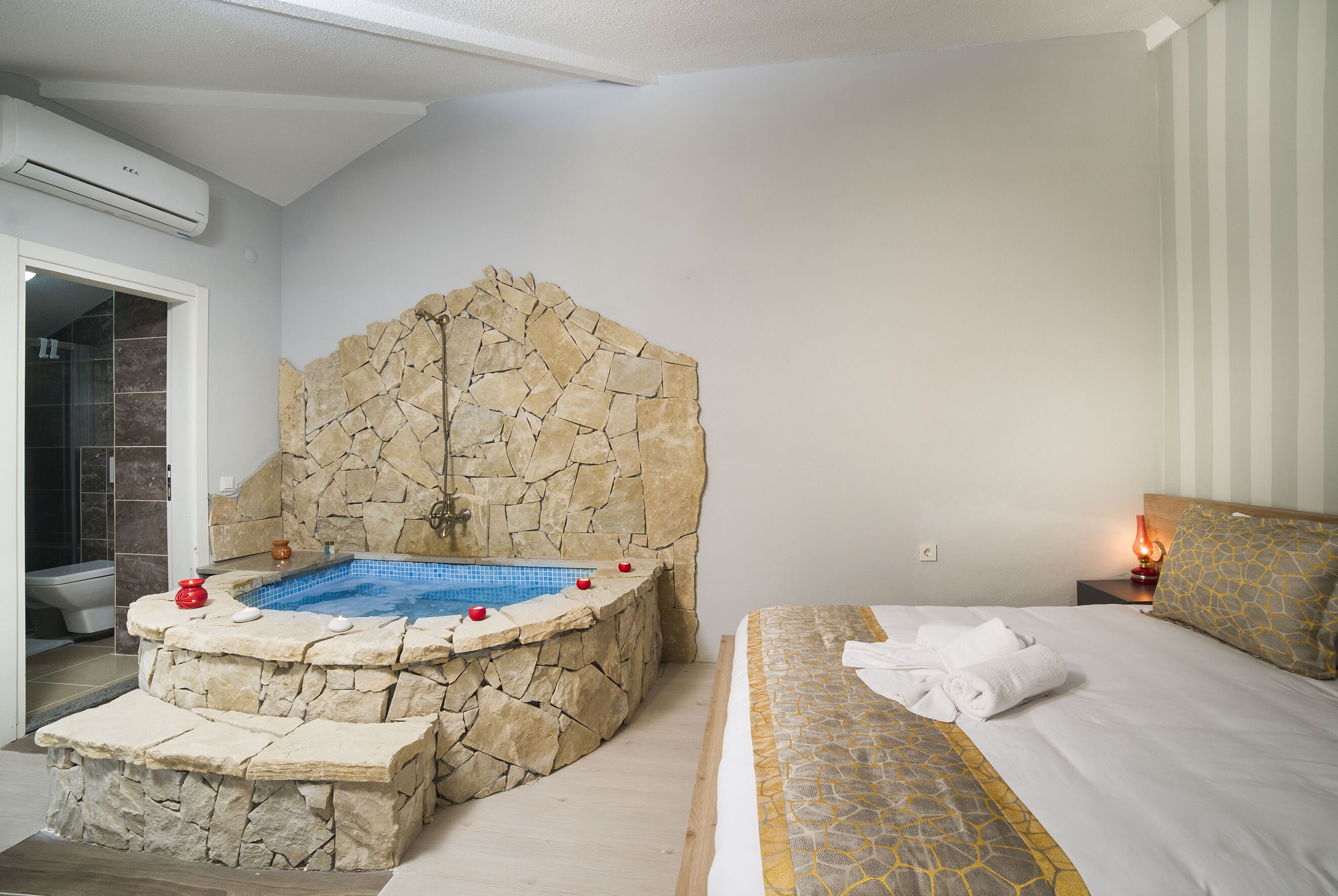 A bedroom with a stone wall, a bed with a patterned bedspread, and a bathroom with a bathtub.