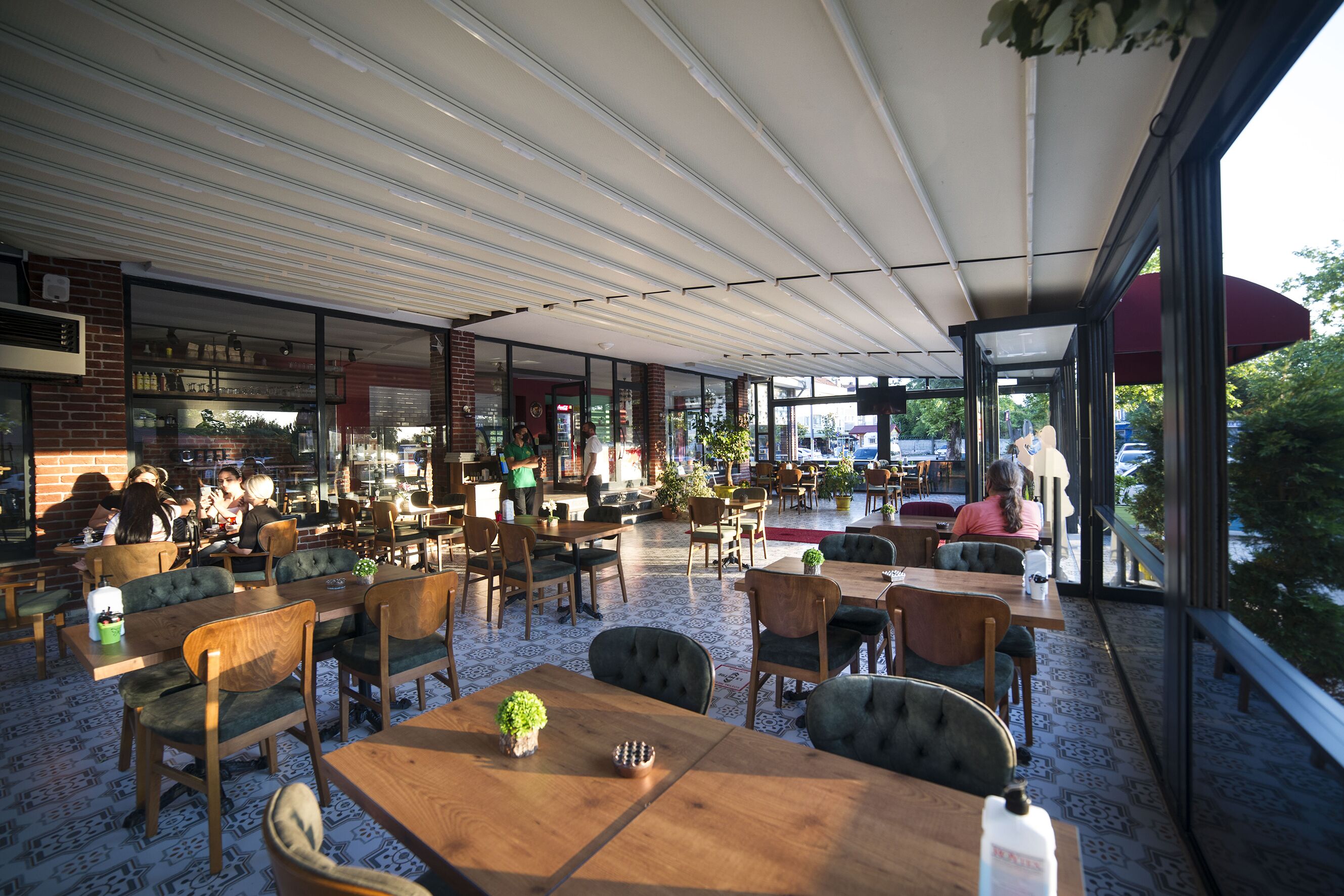 An indoor café with wooden tables and chairs, a brick wall, and large windows offering a view of the outdoors.