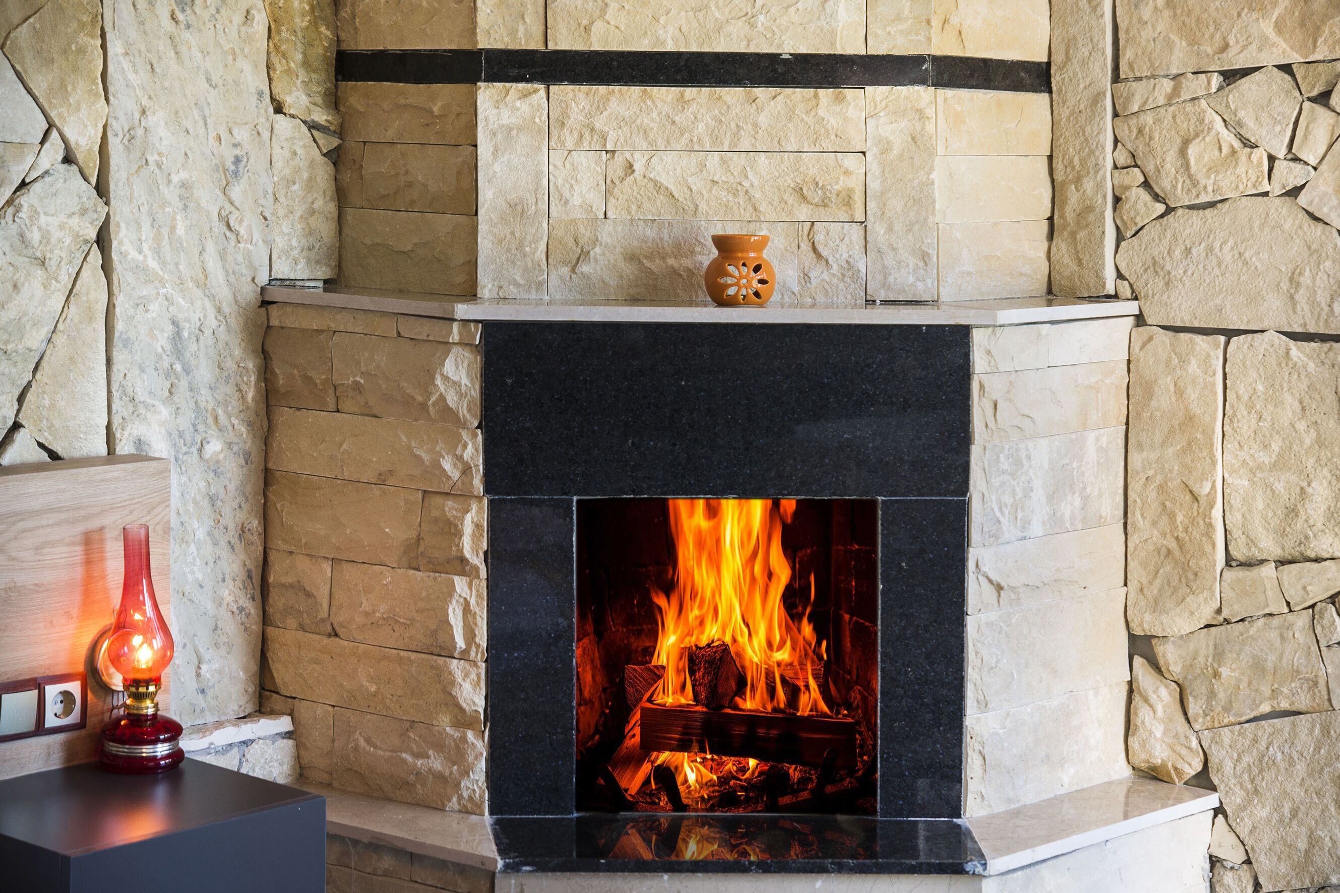 Une cheminée en pierre avec un feu qui crépite à l’intérieur, un vase posé sur le manteau de la cheminée et une bougie sur une table, à gauche.