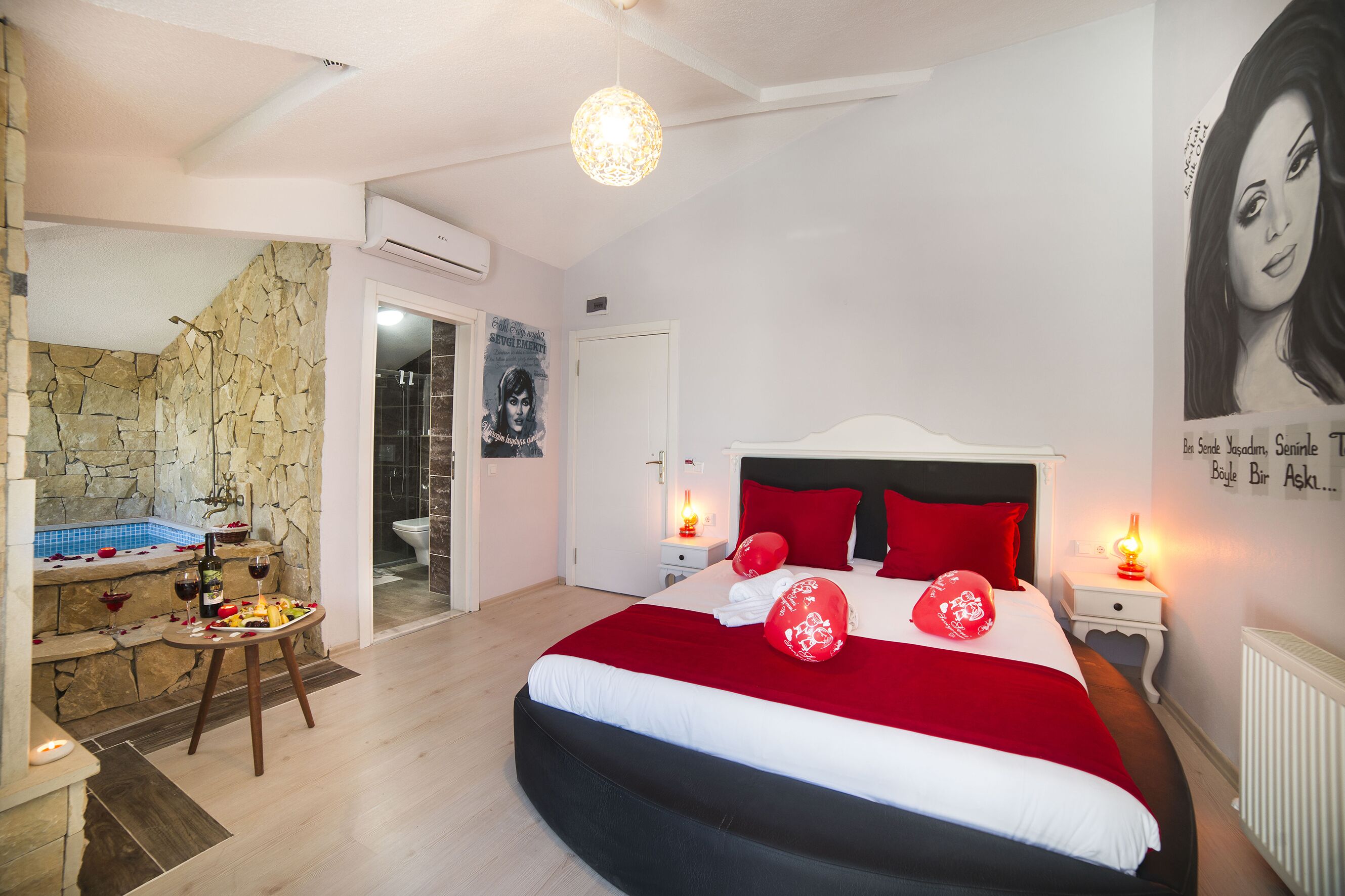 A bedroom with a large bed, a small table with a bottle and glasses, a stone wall, and a bathroom visible through an open door.