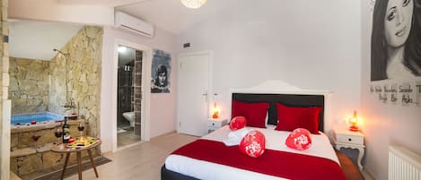 A bedroom with a large bed, a small table with a bottle and glasses, a stone wall, and a bathroom visible through an open door.