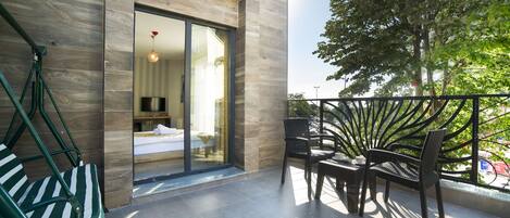 A modern balcony with a glass door leading to a bedroom, a dining area with chairs, and a large tree providing shade.