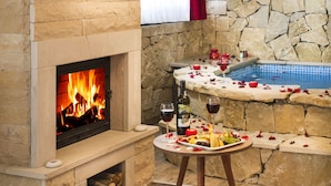A stone fireplace with a fire, a pool area with rose petals, and a table with a bottle of wine and fruit.