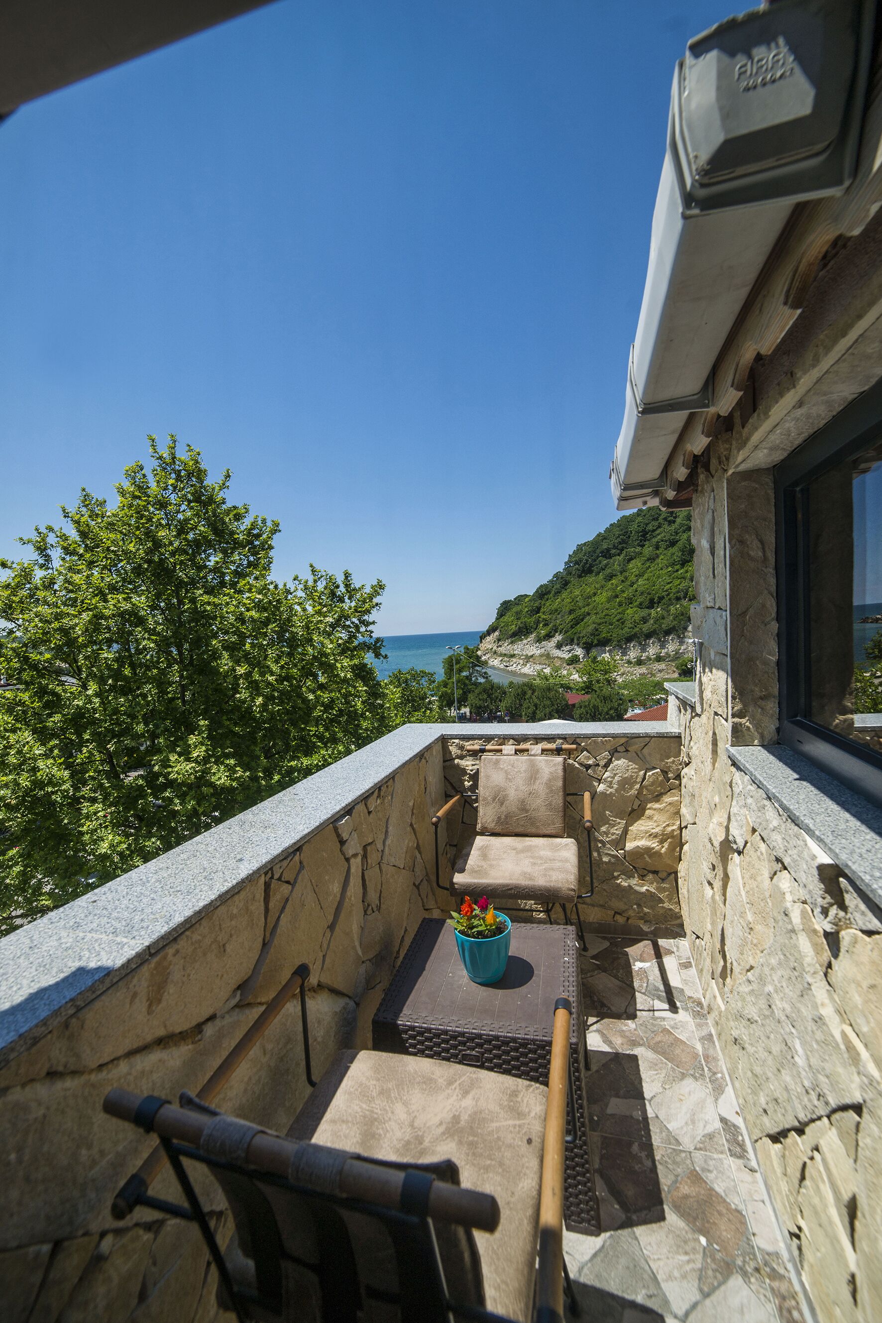 A stone balcony with a view of the sea, a wooden chair, a table, and a potted plant.