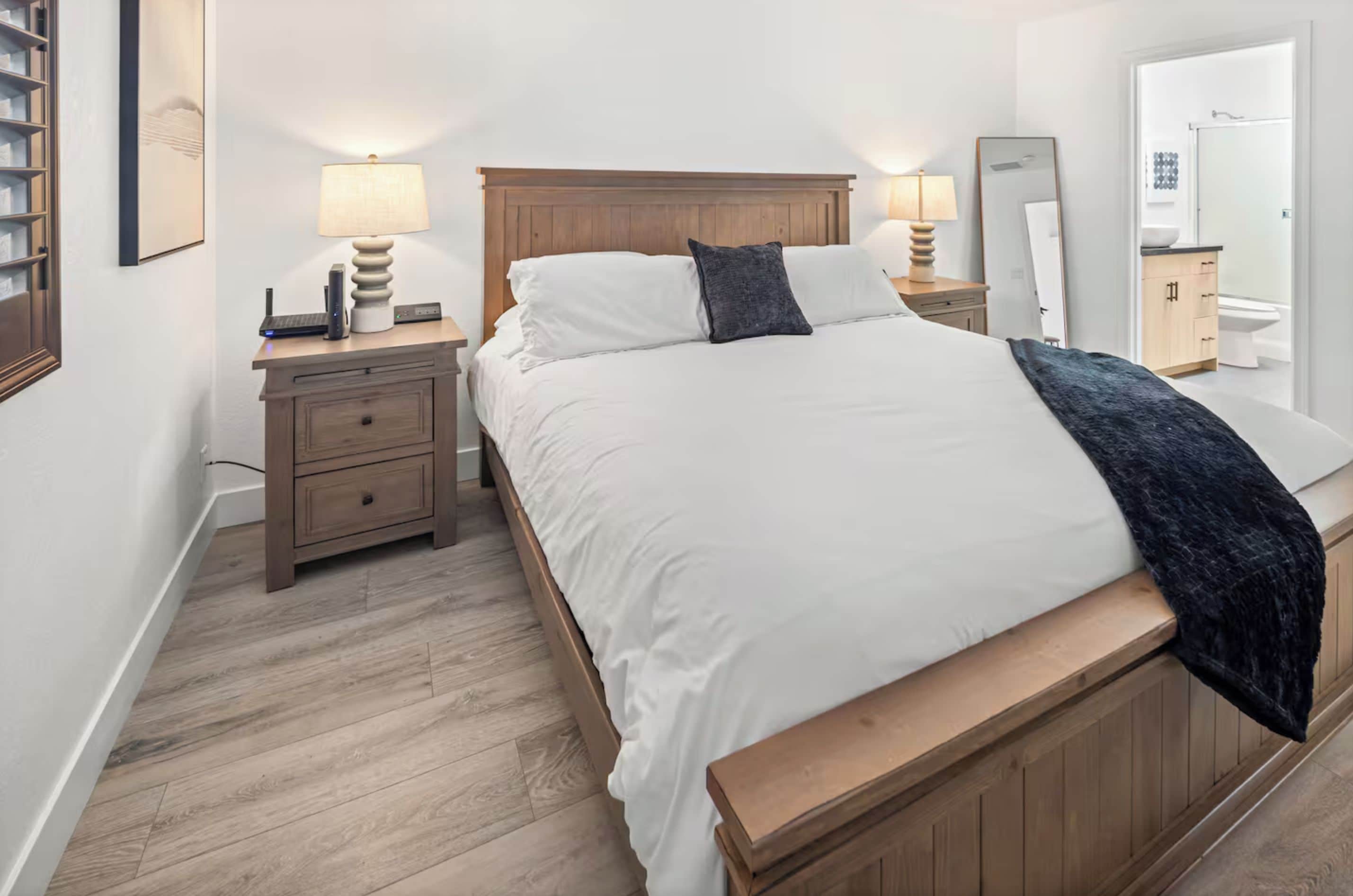 Een slaapkamer met een houten bed, wit beddengoed, een nachtkastje met een lamp en een spiegel.