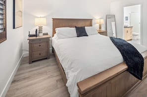 A bedroom with a wooden bed, white bedding, a bedside table with a lamp, and a mirror.
