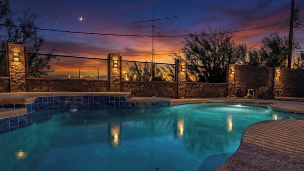 Une piscine avec un mur en pierre et des éclairages, entourée d’arbres et baignée par la lumière d’un coucher de soleil.