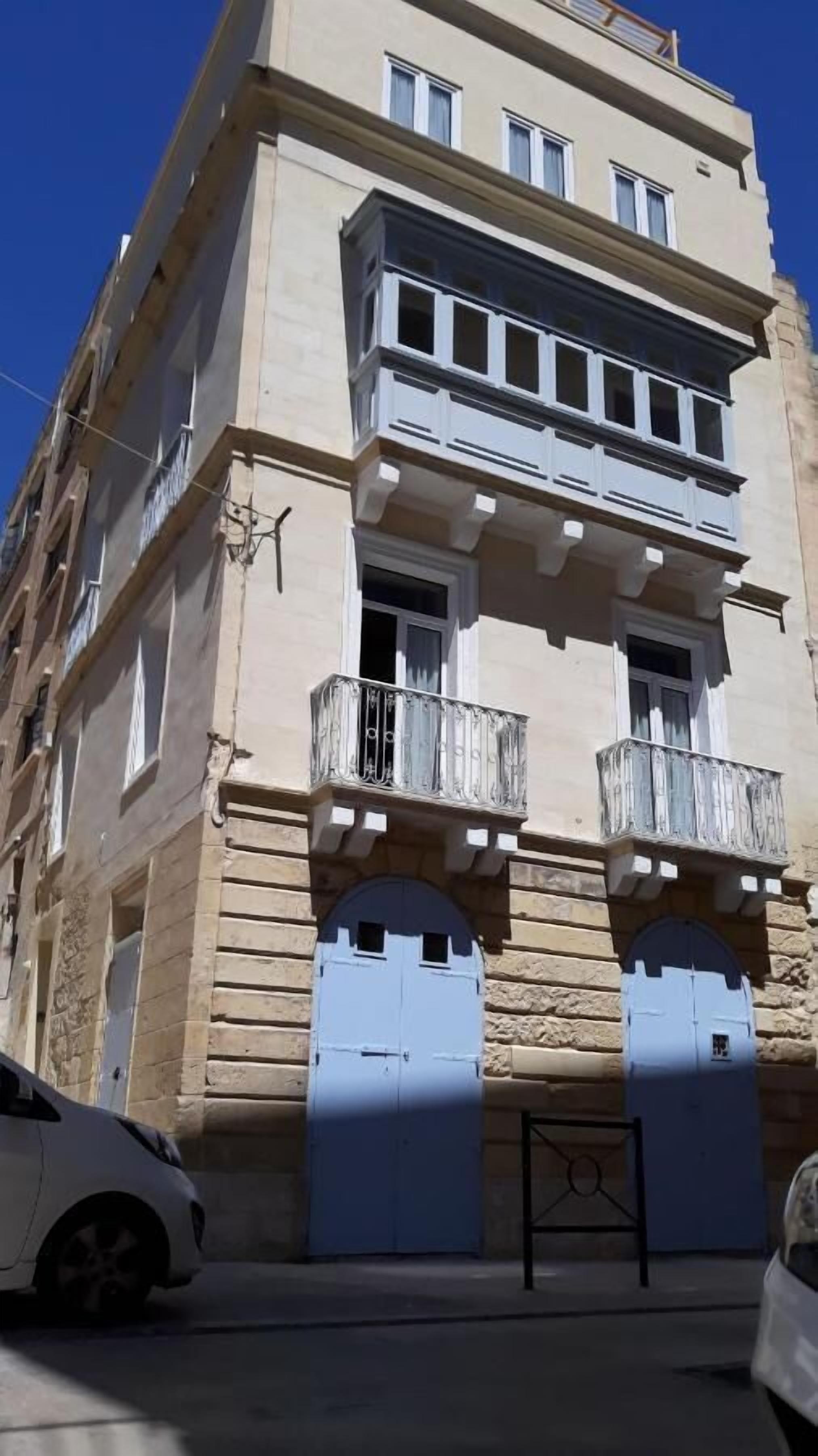 Ein mehrstöckiges Gebäude mit einer beigefarbenen Fassade, zwei Balkonen mit Metallgeländern und einem Paar blaue Doppeltüren.