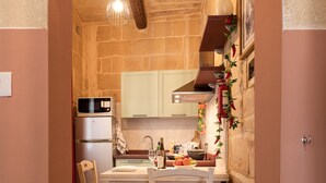 A kitchen with a stone wall, a wooden ceiling, and a tiled floor. There is a white table with chairs, a refrigerator, and a stove. - Concetta Studio Apartments  (Cospicua)