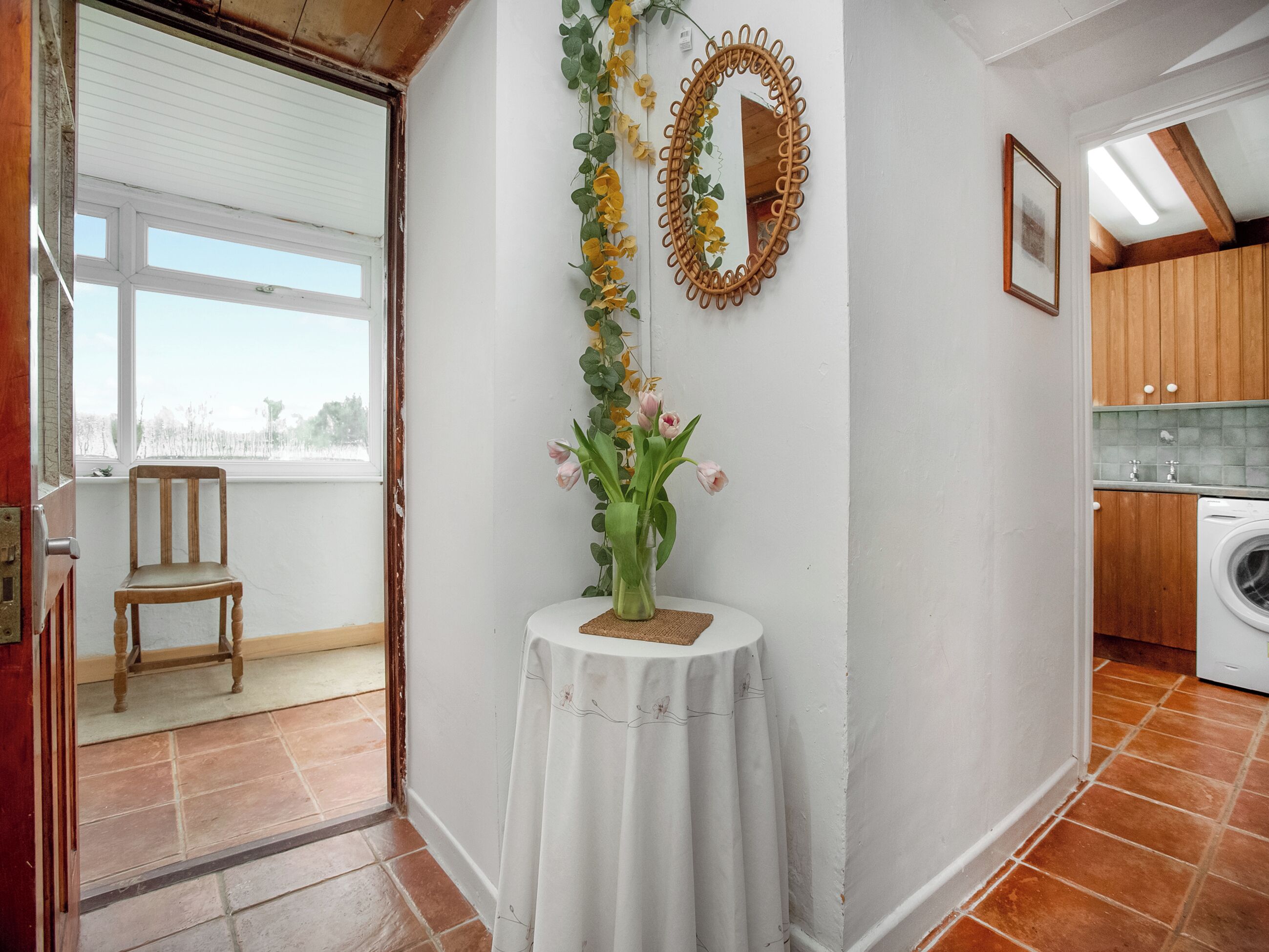 Une pièce avec une chaise en bois près d’une fenêtre, une table avec un vase de fleurs, un miroir et un lave-linge dans un coin.