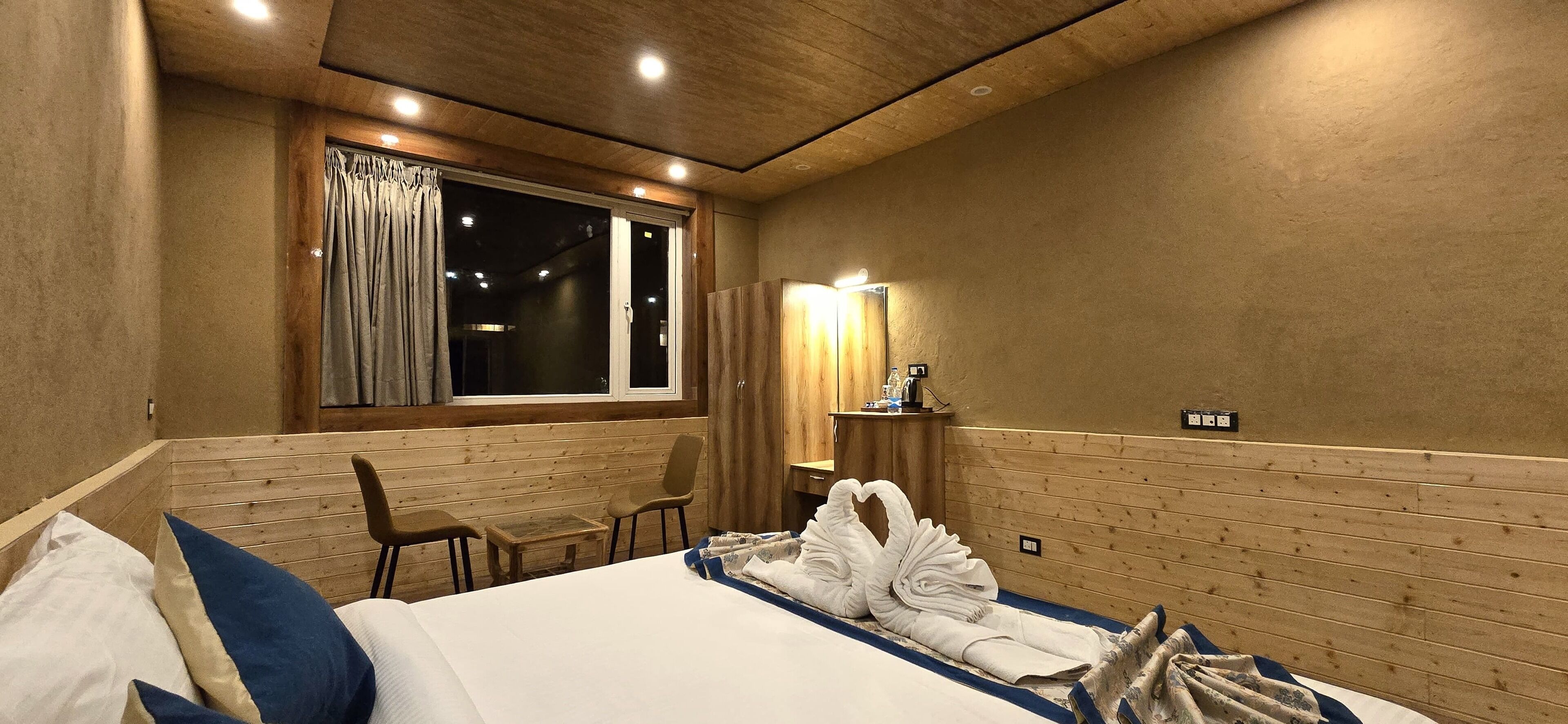 A hotel room with a wooden headboard, a bed with white and blue bedding, a small table, a chair, a window with curtains, and a wall-mounted mirror.