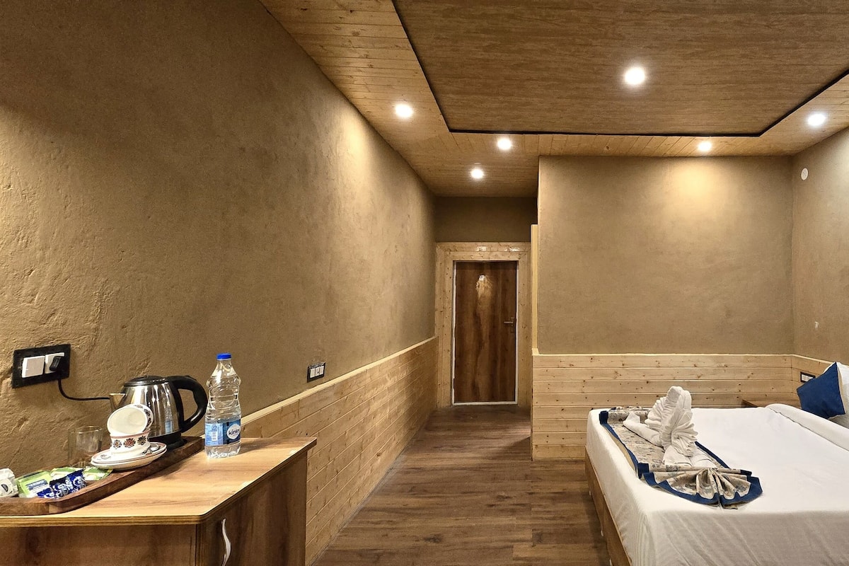 A hotel room with a wooden floor, a bed with white bedding and blue pillows, a bedside table with a kettle and a bottle of water, and a wooden bench along the wall.
