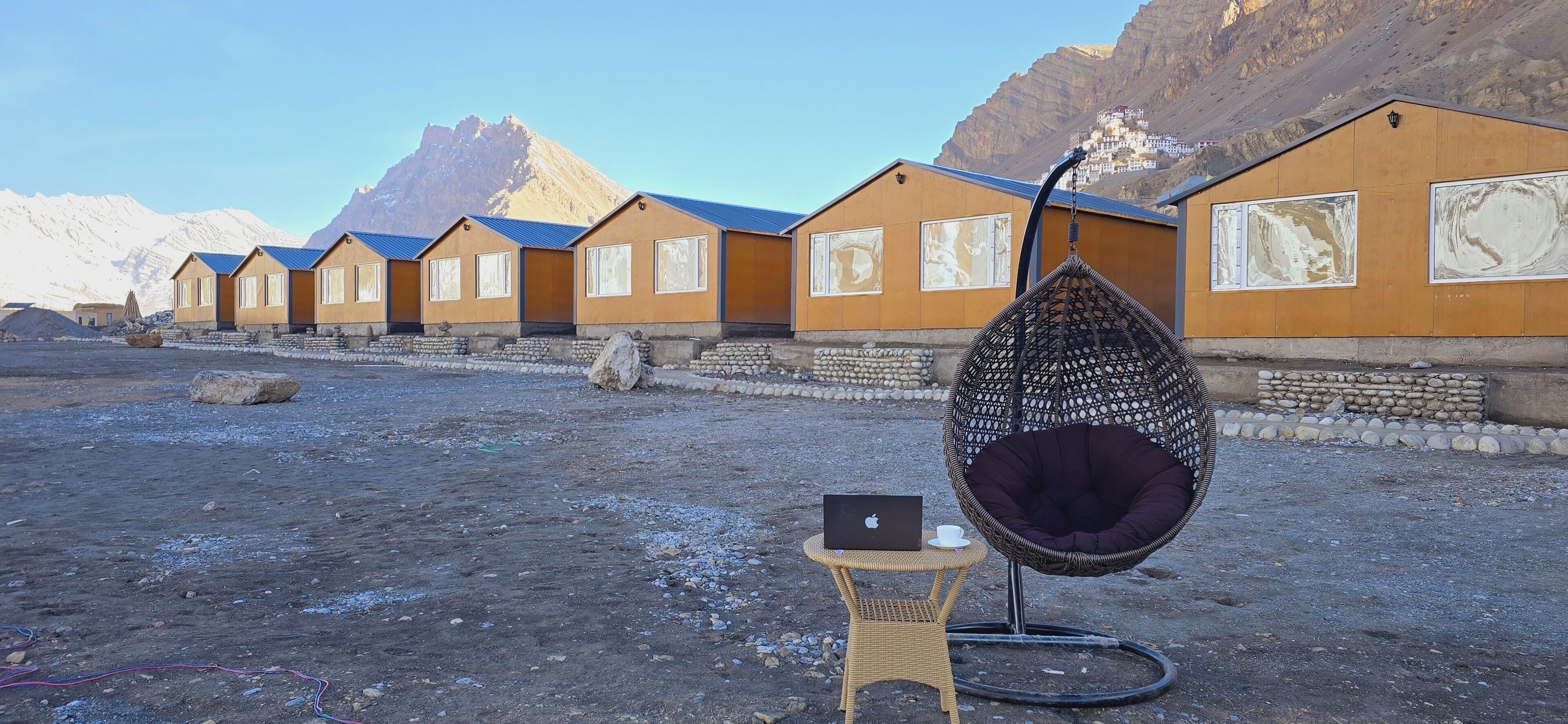 Una hilera de edificios amarillos con techos azules, una silla colgante con cojín, un ordenador portátil sobre una mesita y un paisaje montañoso al fondo.