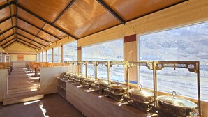 A long, open dining area with wooden benches and tables, large windows with a view of a mountainous landscape, and a series of gold-colored food service stations along the windows. - Hotel Spiti Villa Himalayan Brothers (Spiti)