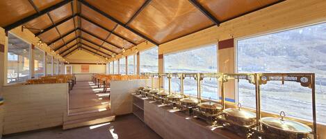 A long, open dining area with wooden benches and tables, large windows with a view of a mountainous landscape, and a series of gold-colored food service stations along the windows.