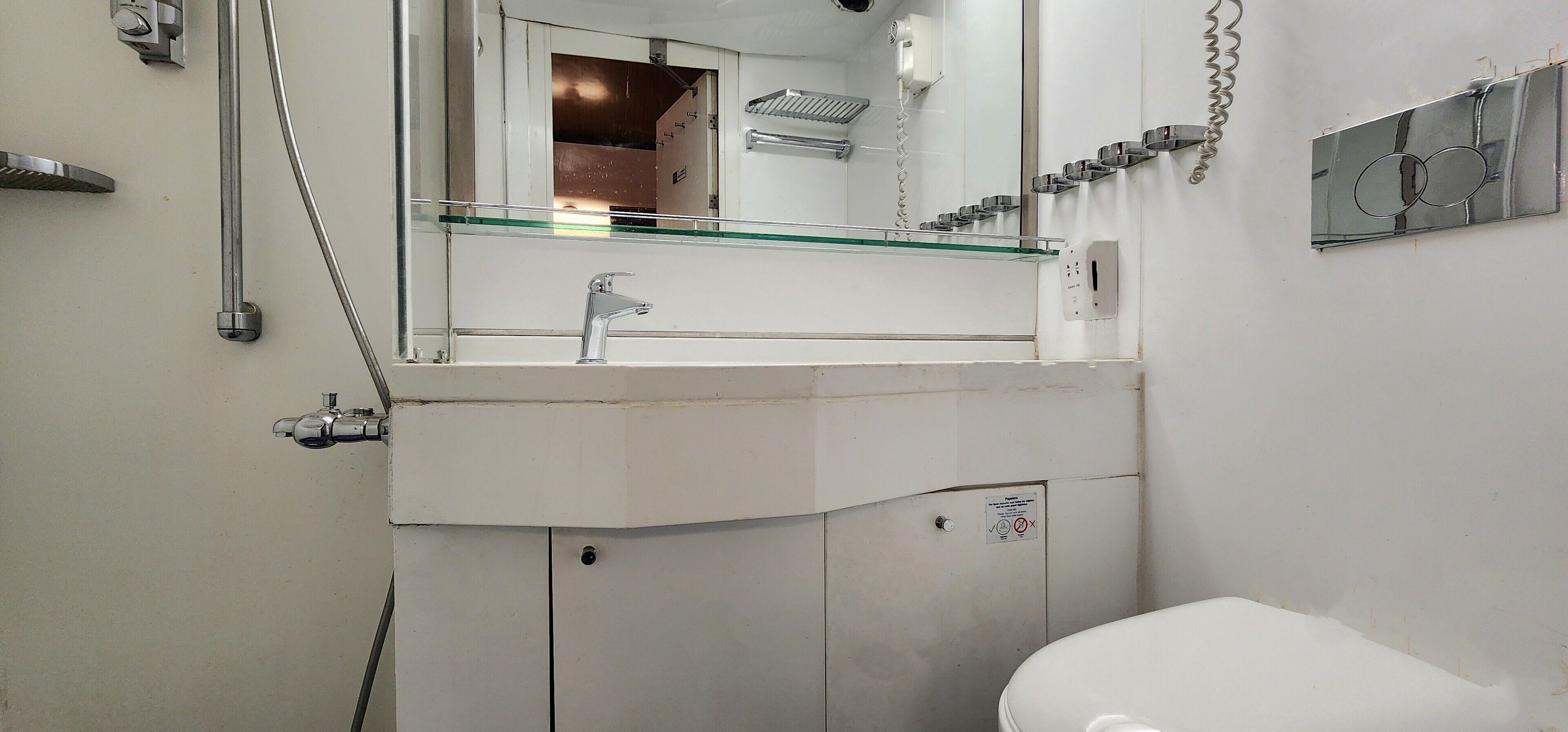 Et badeværelse med et hvidt toilet, en hylde af glas over vasken, og et spejl der afbilder et bruseområde.
