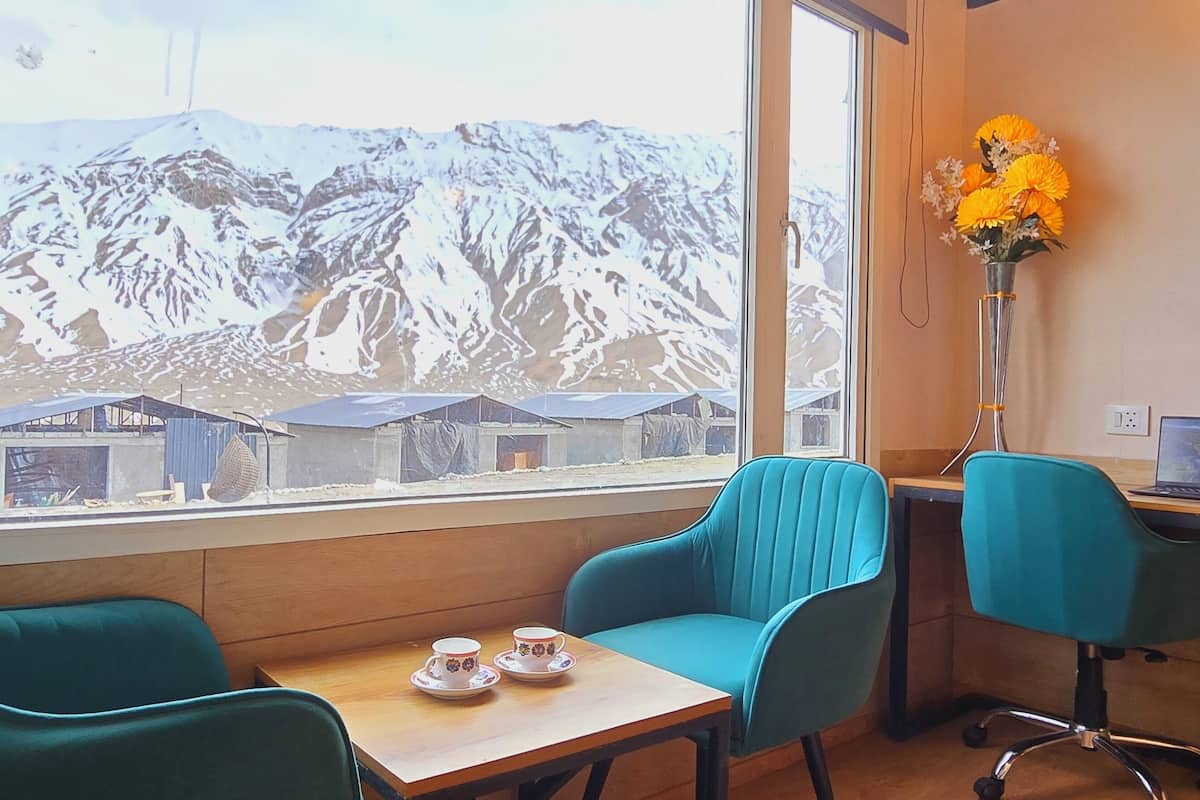 A room with a large window offering a view of snow-covered mountains, two teal chairs, a small table with two cups, and a vase with yellow flowers.