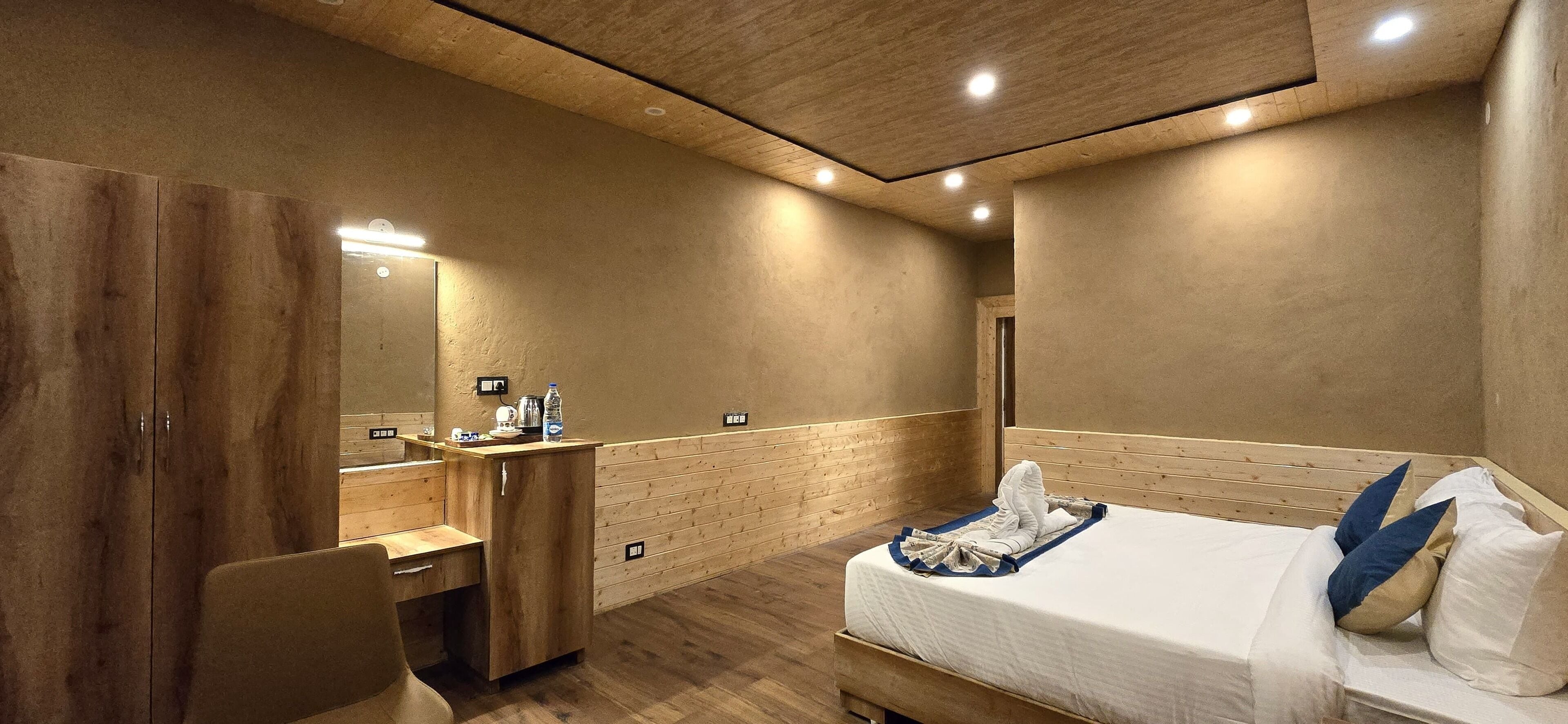 Ett modernt hotellrum med en garderob i trä, ett skrivbord med en spegel, en säng med en blå och vit kudde och ett trägolv.