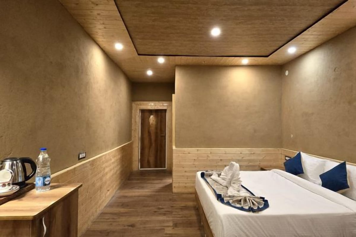 A hotel room with a wooden floor, a bed with white bedding and blue pillows, a wooden desk with a kettle and water bottle, and a wooden door leading to another room.