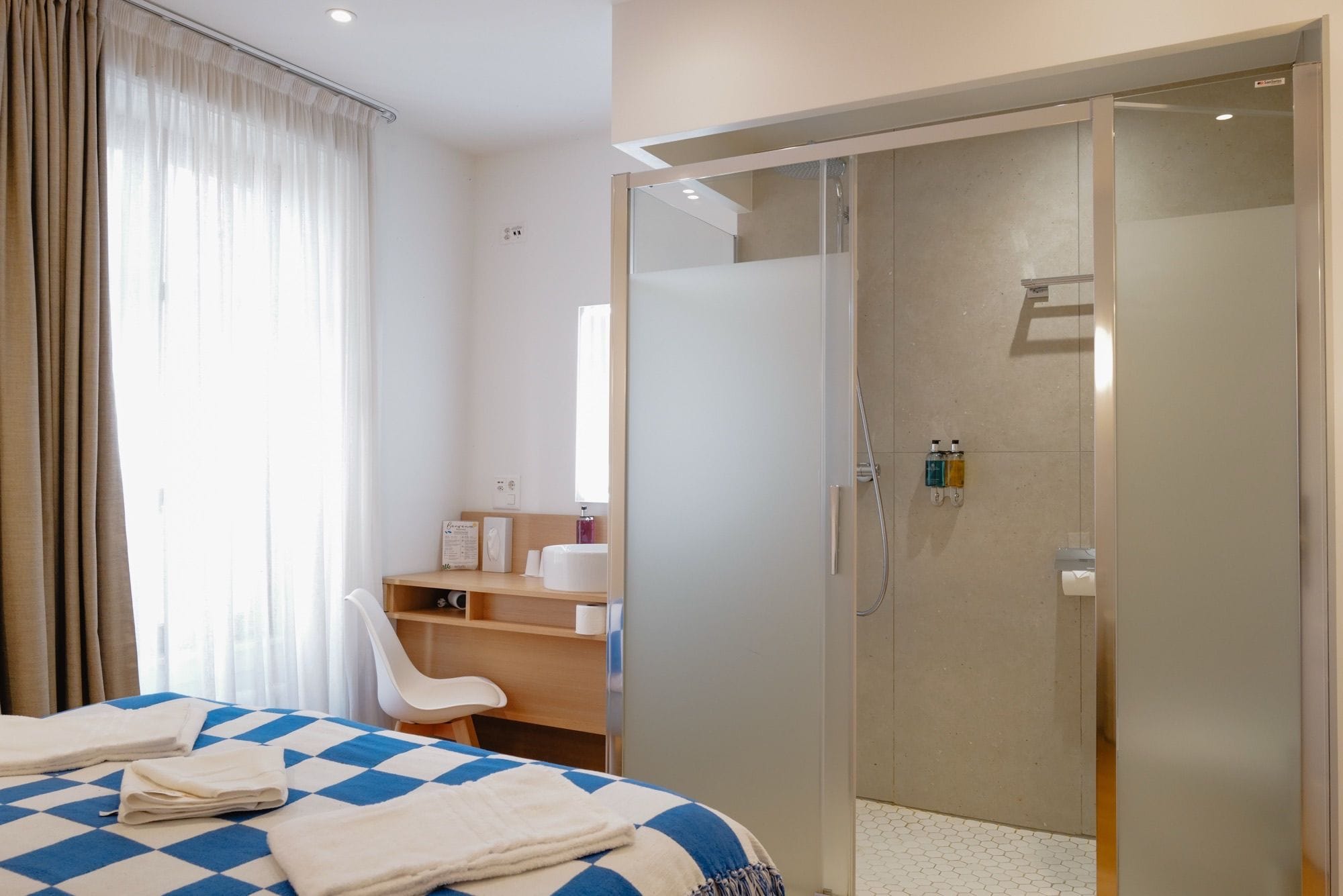 A hotel room with a bed, a chair, a desk, a wardrobe, and a shower area.