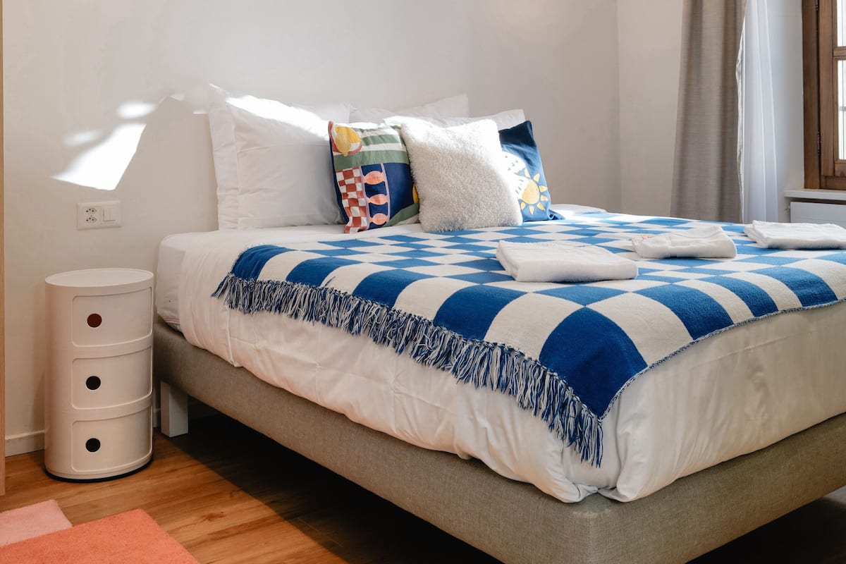 A bedroom with a bed, bedside tables, a nightstand, a window with curtains, and a wall-mounted light fixture.