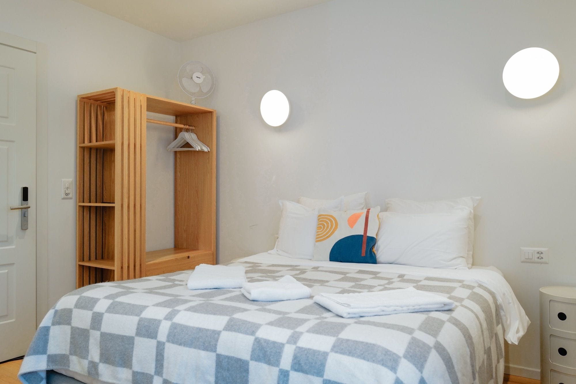A hotel room with a bed, a wooden wardrobe, a wall-mounted fan, and a bedside table.