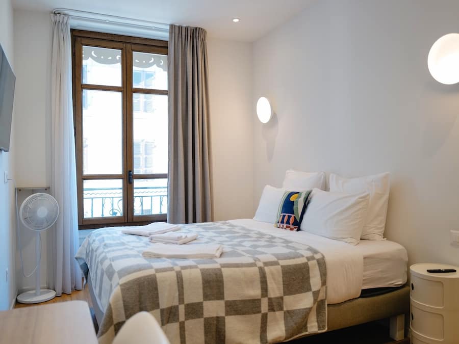 Una habitación de hotel con cama, televisor montado en la pared, ventilador de pie, ventana con cortinas y mesita de noche con lámpara.