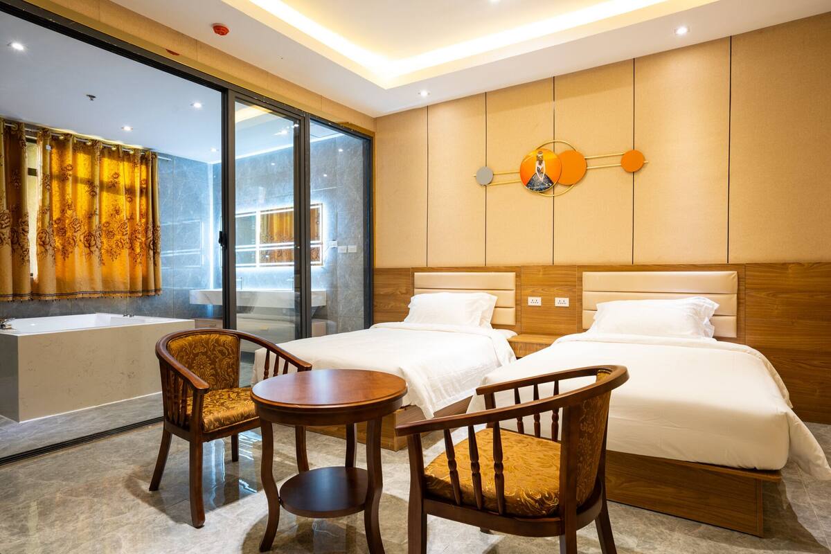 A hotel room with two beds, a sitting area with a table and chairs, a large window with curtains, and a wall-mounted light fixture.